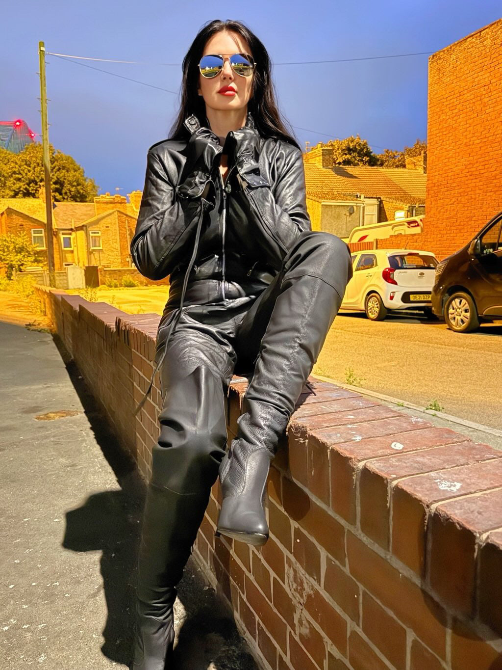 Woman in black leather outfit sitting on a brick wall with a street and cars in the background
